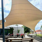 Outdoor dining area with wooden picnic tables under a large beige shade sail, metal posts, and potted flowers.
