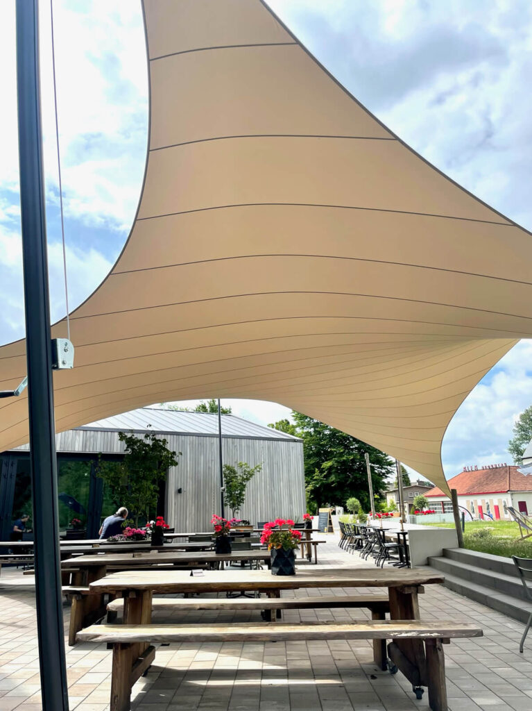 Outdoor dining area with wooden picnic tables under a large beige shade sail, metal posts, and potted flowers.