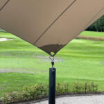 Shade sail over a golf course putting green, with a dark green pole and distant flag markers on the fairway