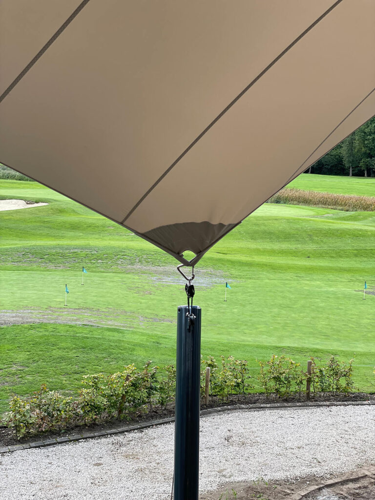 Shade sail over a golf course putting green, with a dark green pole and distant flag markers on the fairway