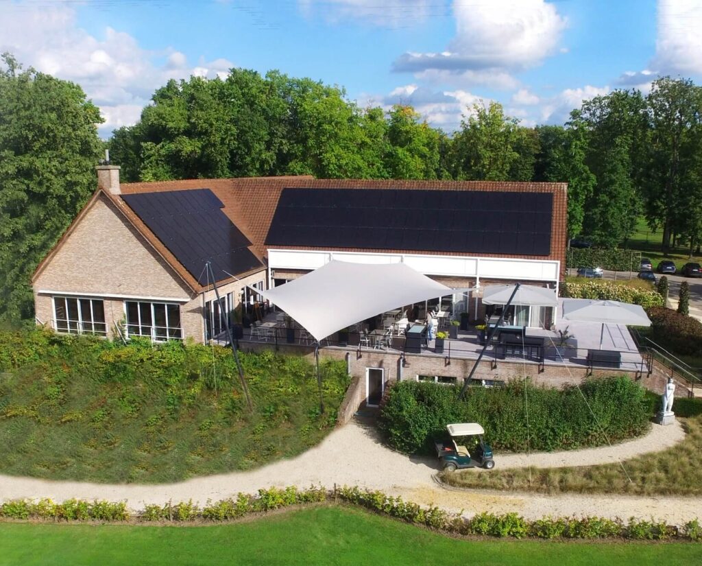 Brick clubhouse with dark solar panels on the roof; outdoor patio with white shade sails and seating among trees.