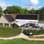 Brick clubhouse with dark solar panels on the roof; outdoor patio with white shade sails and seating among trees.