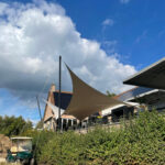 Outdoor area with beige sail canopies over a patio beside a brick building under a blue sky with white clouds.