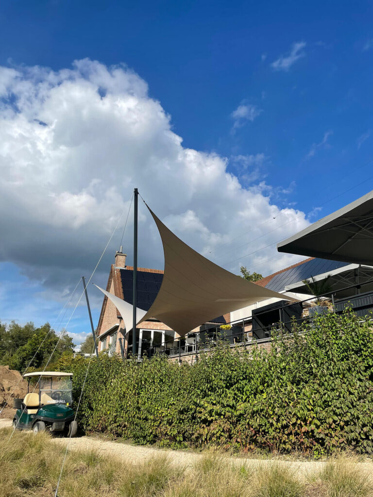Outdoor area with beige sail canopies over a patio beside a brick building under a blue sky with white clouds.