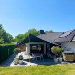 Backyard patio with a black triangular shade sail, white outdoor furniture, potted trees, and a modern house with a tiled roof.
