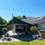 Backyard patio with a black triangular shade sail over a white dining set and potted plants, in front of a tiled-roof house.