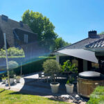 Backyard patio with round wooden hot tub, white potted plants, and dining chairs under a triangular shade sail against a blue sky.