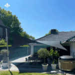 Outdoor patio with a dining table and chairs under black shade sails, surrounded by potted plants and a wooden hot tub beside a tiled house.