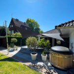 Sunny backyard patio with a large dark shade sail, outdoor dining set, and potted plants under a clear blue sky.