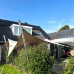 Residential backyard with a beige shade sail, modern house, and solar panels on the roof under a blue sky.