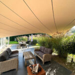 Outdoor patio under a large beige shade sail with wicker couches around a square fire pit and a garden backdrop