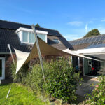Backyard of a house with solar panels on the roof, tall beige shade sails, and lush green plants under a clear blue sky.