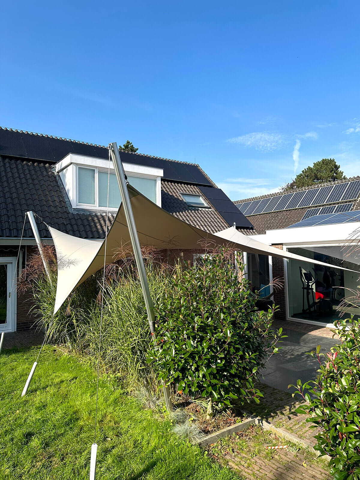 Utrecht-terras-overkapping-op-maat-stormvast-key-doek-maatwerk-waterdicht-lichtdoorlatend-luxe-06 Backyard of a house with solar panels on the roof, tall beige shade sails, and lush green plants under a clear blue sky.