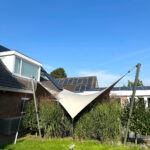 White fabric shade sail stretched between metal poles over a grassy backyard with tall grasses, and solar panels on the house roof in the background.