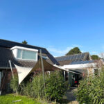 Modern house exterior with solar panels on the roof, a beige triangular shade sail over the patio, and tall grasses in the yard under a clear blue sky.
