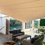 Outdoor patio with a beige sail shade over wicker seating and a rectangular fire pit, beside a modern house with glass doors and a pool visible in the background.
