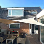Backyard patio next to a brick house with wicker seating, a wooden table, shade sails, and solar panels on the roof.