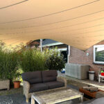 Outdoor patio with gray-cushioned wicker seating under a large beige shade sail; brick house in the background with plants nearby.