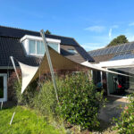 Backyard of a modern house with solar panels on the roof, beige triangular shade sails, and green shrubs in the foreground.