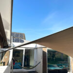 Backyard patio with a hammock, wicker sofa, and a beige sun shade sail stretched above a brick patio, with sliding glass doors to the house and solar panels on the roof in the background.
