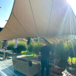 Man standing with back to camera under a large beige shade sail over an outdoor wicker seating area in a sunny backyard.