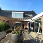 Sunny backyard patio with a weathered wooden table and a large black potted plant, wicker chairs, and beige sail shades; brick house with solar panels on the roof in the background.