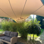 Outdoor seating area under a large beige sun shade with a wicker sofa and surrounding tall grasses and a small tree nearby.