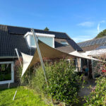 Backyard of a modern house with solar panels on the roof, a beige shade sail supported by a pole, and green lawn with shrubs.