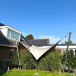 White fabric shade sail stretched over a grassy backyard next to a house with solar panels on the roof, under a clear blue sky.