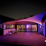 Nighttime terrace of a modern building bathed in purple and pink lighting from curved fabric canopies, glass doors reveal an interior lounge area; logo in corner.