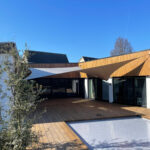 Modern house exterior with wooden deck, large glass doors, and white fabric shade sails against a clear blue sky