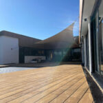 Wooden deck patio beside a modern building with a beige fabric shade sail and clear blue sky.