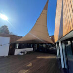 Beige fabric sun shade stretched between a tall pole and a modern wooden building over a wooden deck courtyard.