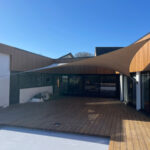 Open-air wooden deck courtyard with a large beige fabric shade sail spanning between the building and a white wall, in front of glass doors.