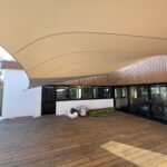 Large beige tensile canopy over a wooden deck outside a modern building with glass doors and windows.