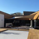 Modern courtyard/patio with wooden decking, glass walls, and white pool cover under cream fabric shade sails.