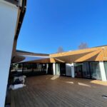 Wooden deck courtyard of a modern building with beige fabric shade sails and glass doors beyond.