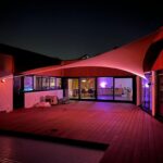 Nighttime rooftop patio with a curved tensile canopy lit in pink, glass doors to interior, seating visible inside.
