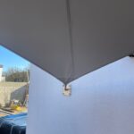 Gray outdoor shade sail attached to a light blue wall with a metal mounting bracket and tension cable running along its edge.