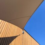 Beige shade sail above a vertical wooden wall, set against a bright blue sky and sunny daylight.