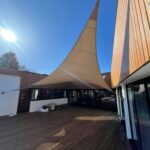 Beige tensile shade sail stretched between a building and posts over a wooden deck courtyard.