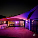 Nighttime terrace of a modern home with a large pink-purple fabric shade sail, glowing interior lights visible through glass doors.