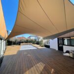 Wooden patio deck with a large beige fabric shade sail stretched above, blue sky visible between buildings.