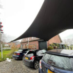 Large black fabric shade sail stretching over a gravel parking area with several cars and brick houses in the background, providing shelter from sun.