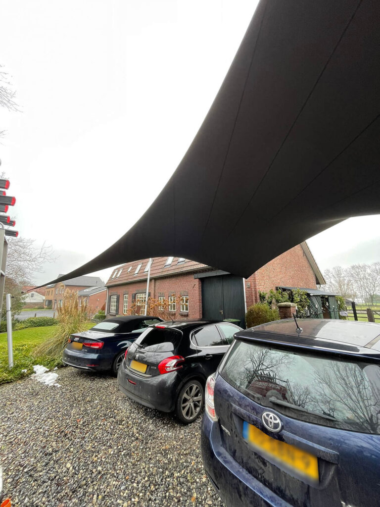 Large black fabric shade sail stretching over a gravel parking area with several cars and brick houses in the background, providing shelter from sun.