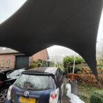 Large curved black awning or sail hanging over a parked blue Toyota Verso in a driveway.