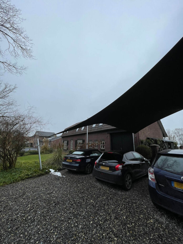 Parking lot with three cars under a large diagonal black canopy, brick building in the background and leafless trees on the left.