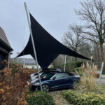 Dark triangular shade sail stretched between metal poles above a blue convertible parked in a residential driveway, with bare trees and garden shrubs nearby.