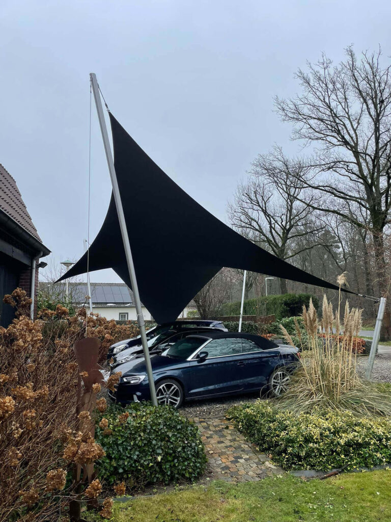 Dark triangular shade sail stretched between metal poles above a blue convertible parked in a residential driveway, with bare trees and garden shrubs nearby.