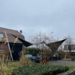 Black triangular shade sails over a driveway with cars, a brick house, and bare trees on a cloudy day.